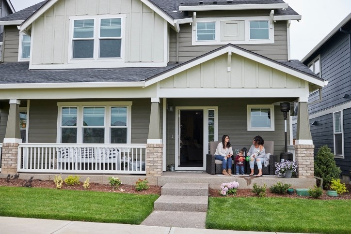 Two women, a child, and a small white dog sit on the front porch of a new home in Reed’s Crossing.